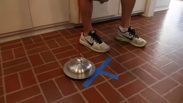 Silver sensor disc standing on a red tile floor beside a blue tape cross, with a person's sneakers behind it