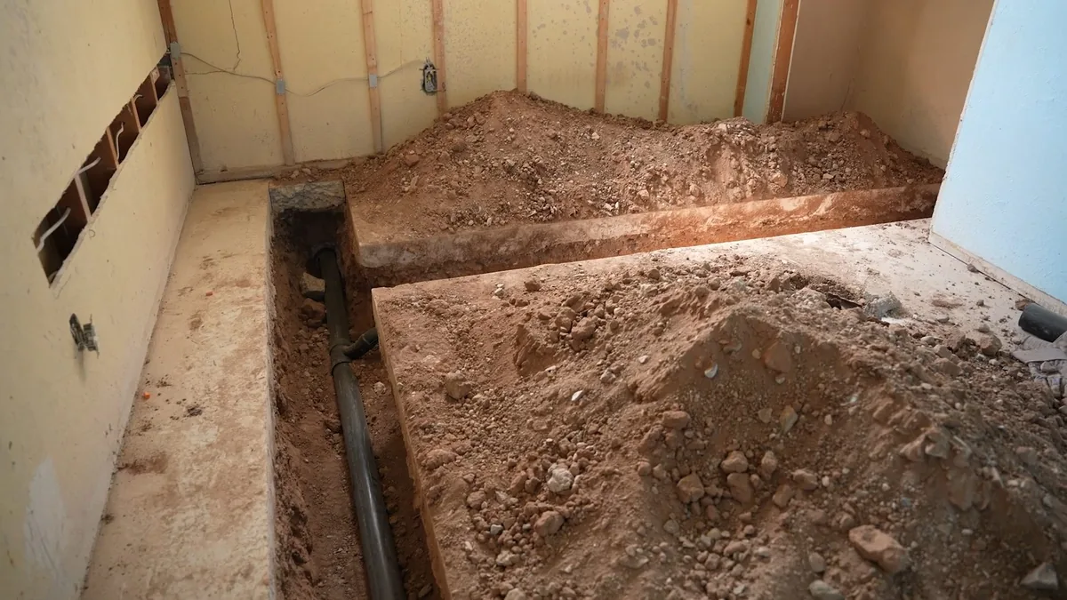 Basement room with a trench dug through the floor along a wall, soil piled either side and a pipe lying in the bottom