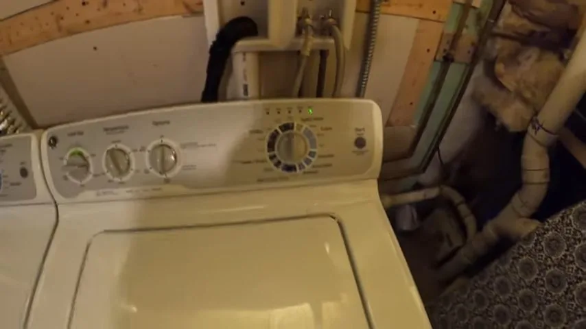 Cream top-load washer in a basement with pipes and a drain hose rising behind it along a wall