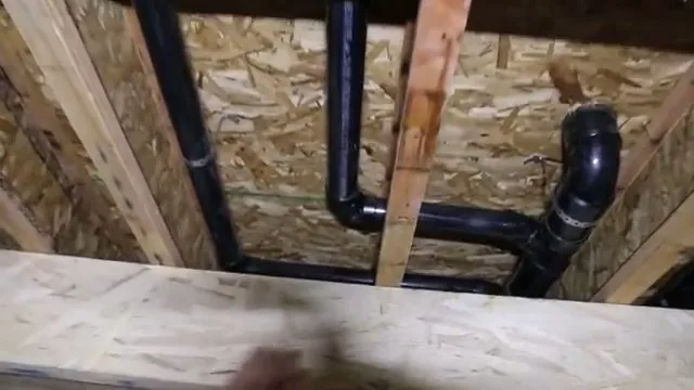 Black plastic drain pipes and a trap running between wood joists above a wood subfloor