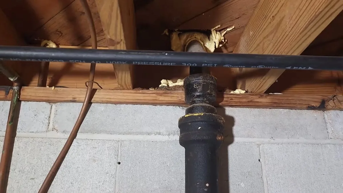 Black cast iron drain stack rising through a wood joist above a concrete block wall, with a black flexible pipe and a copper line beside it
