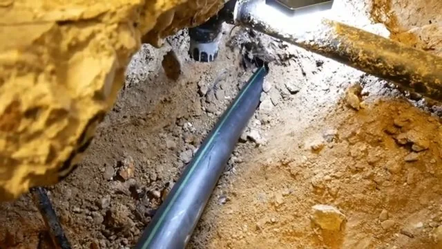 Black drain pipe lying in orange clay soil in a dug trench beneath a rock overhang, lit by a work light
