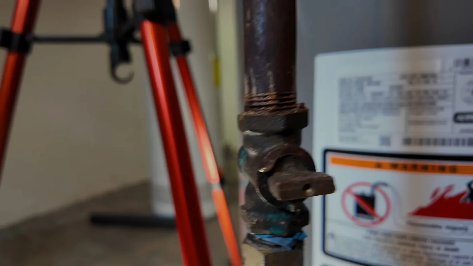 Black iron gas shutoff valve beside a water heater, with a warning label and a red tripod behind it