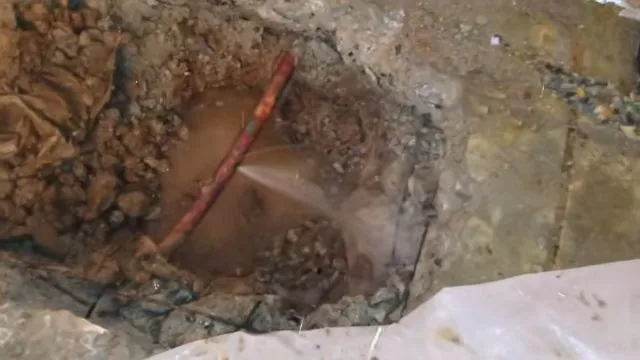 Muddy hole in a floor with a copper line spraying water across the pooled water in it
