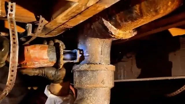 Corroded cast iron drain joint under wood floor joists, with a worker's hand and a power saw at the joint