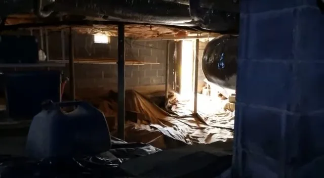 Dark crawlspace under a house with plastic sheeting on the ground, a block foundation wall and a plastic jug