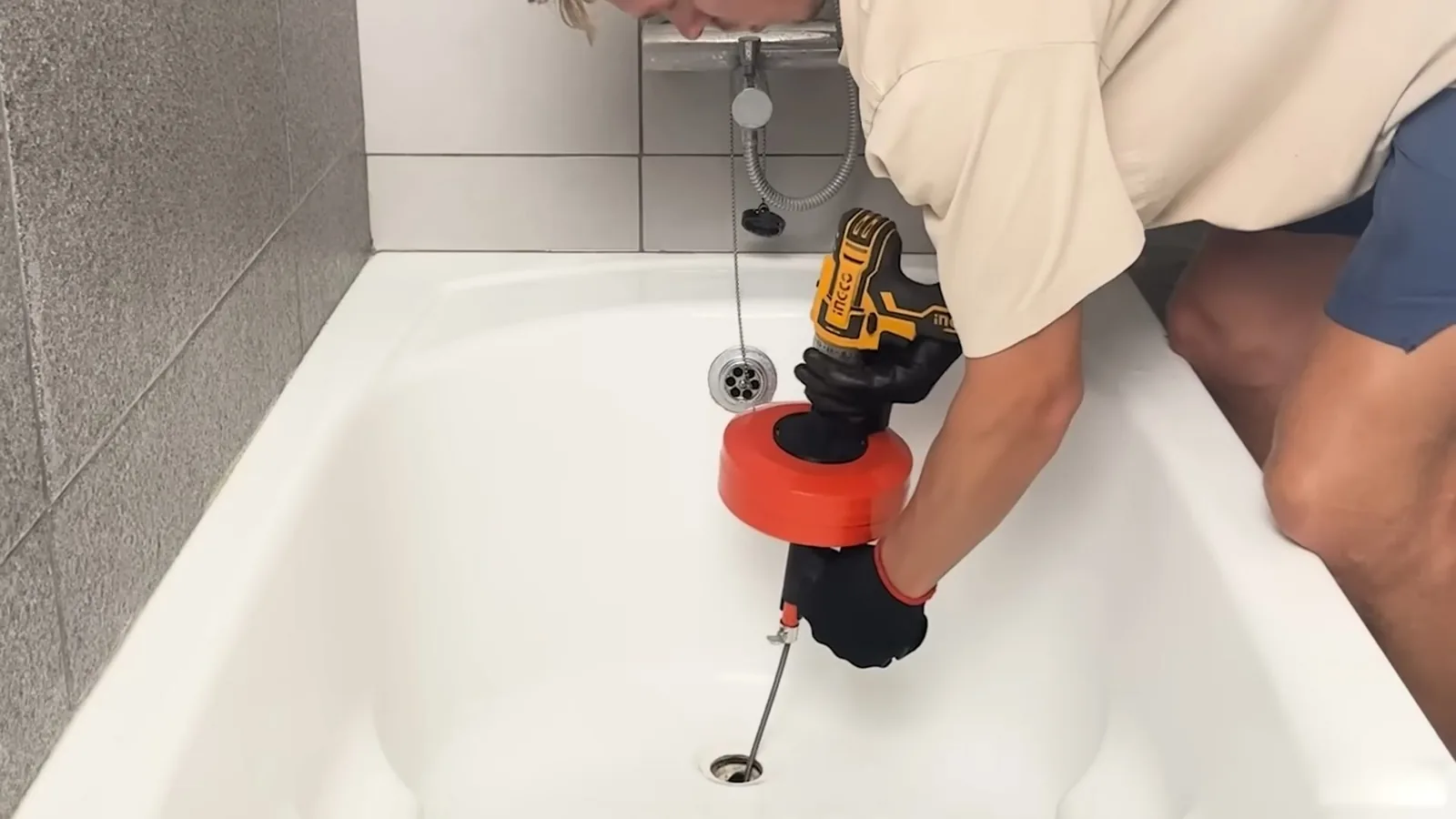 Person bending over a white bathtub to run a cordless drill and drain snake down the drain