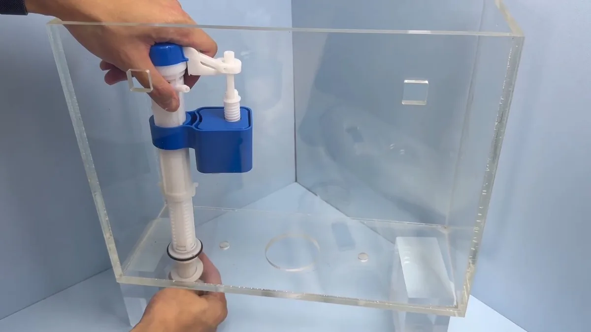 Toilet fill valve and flush valve assembly inside a clear plastic tank, with a hand reaching in