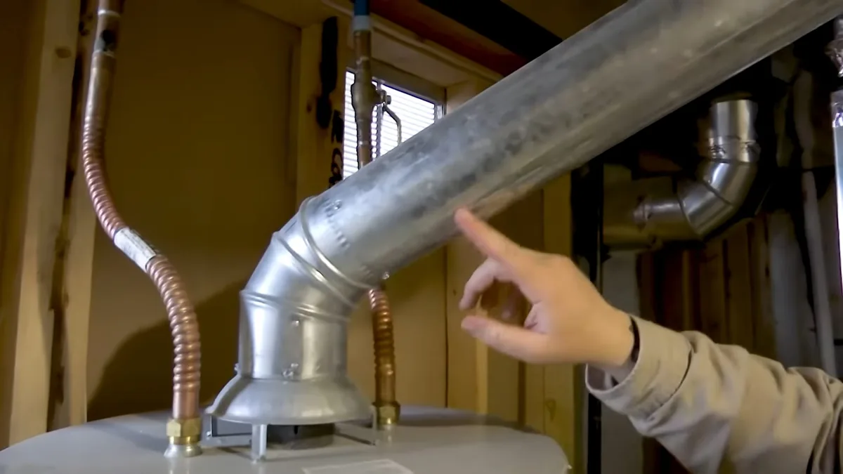Hand pointing at the metal flue vent connection on top of a gas water heater, with copper lines beside it