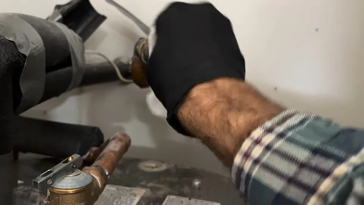 Gloved hand in a plaid sleeve reaching to a hose connection on top of a water heater, beside a relief valve