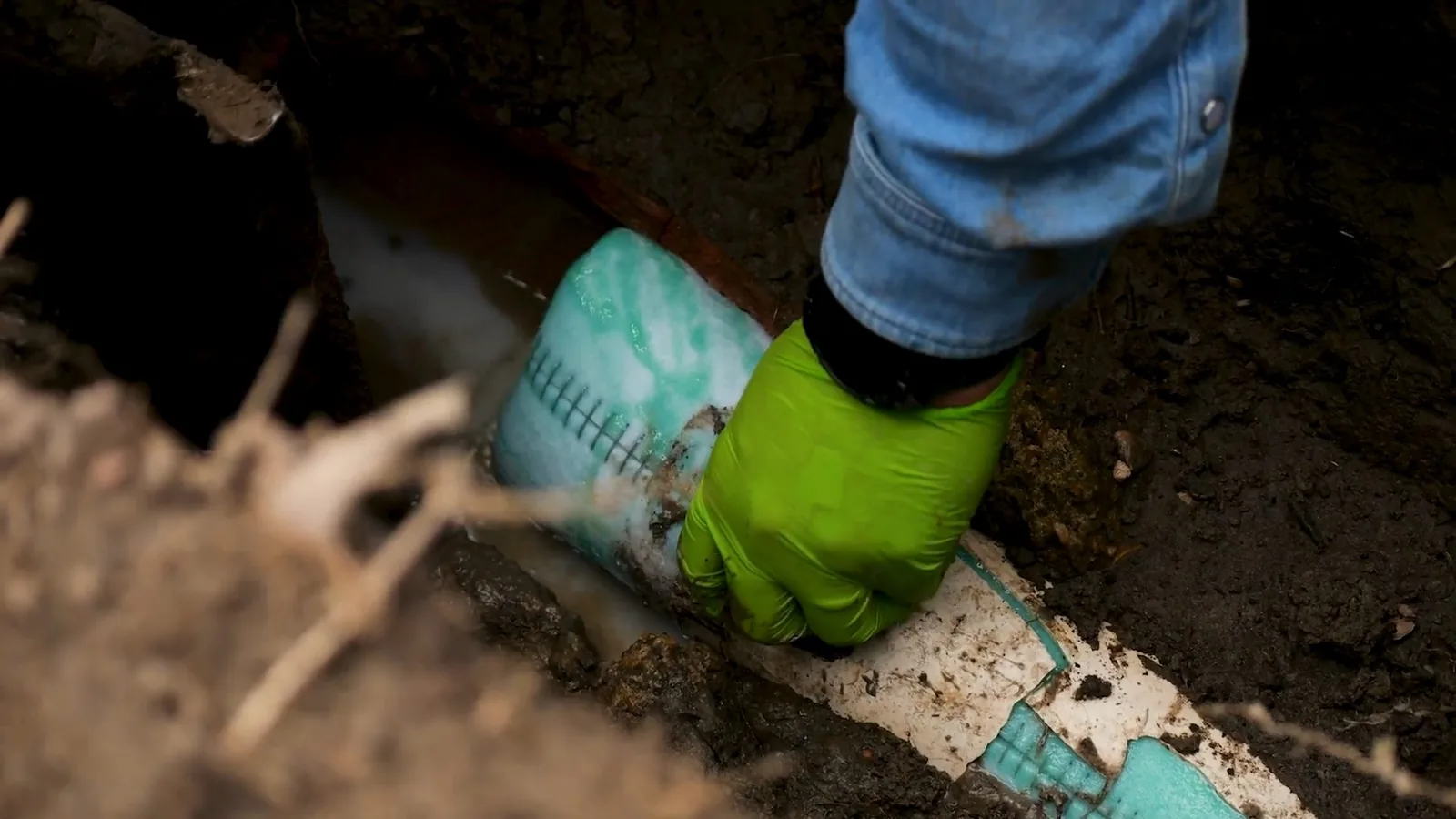 Gloved hand guiding a green pipe liner sleeve into a pipe in a dirt excavation