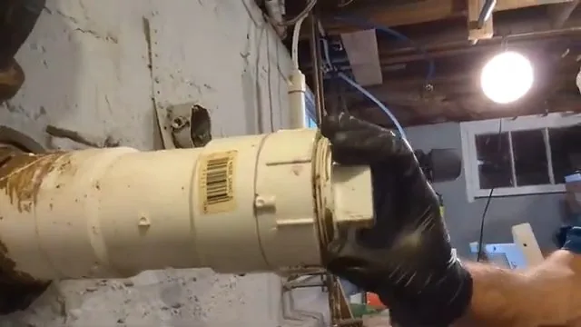 Black-gloved hand gripping a cream-coloured plastic cleanout fitting on a drain line in an unfinished basement
