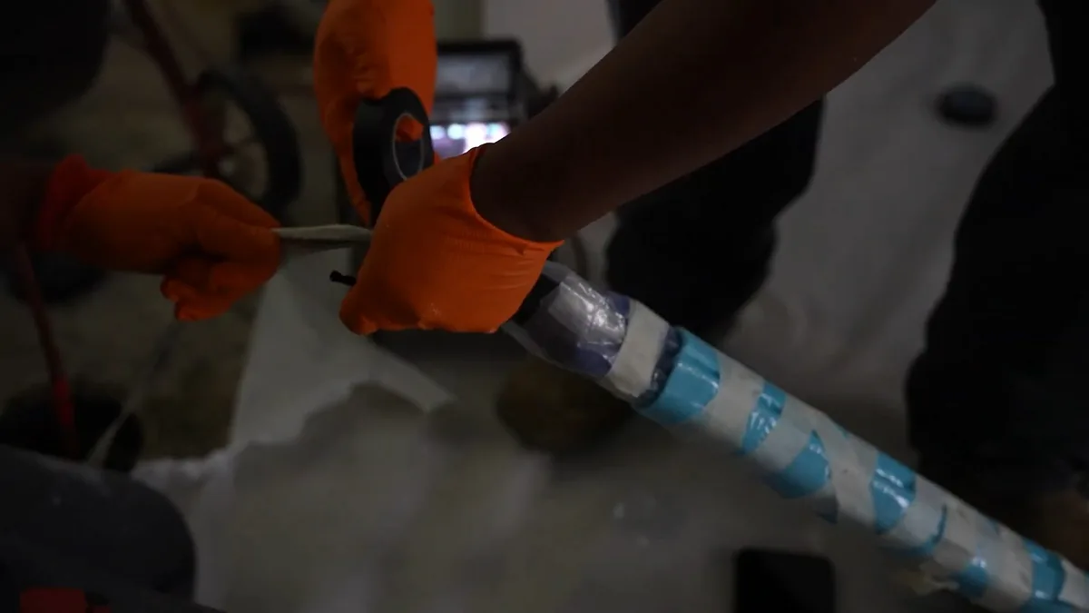 Orange-gloved hands wrapping tape around a pipe liner covered in blue and white spiral wrap