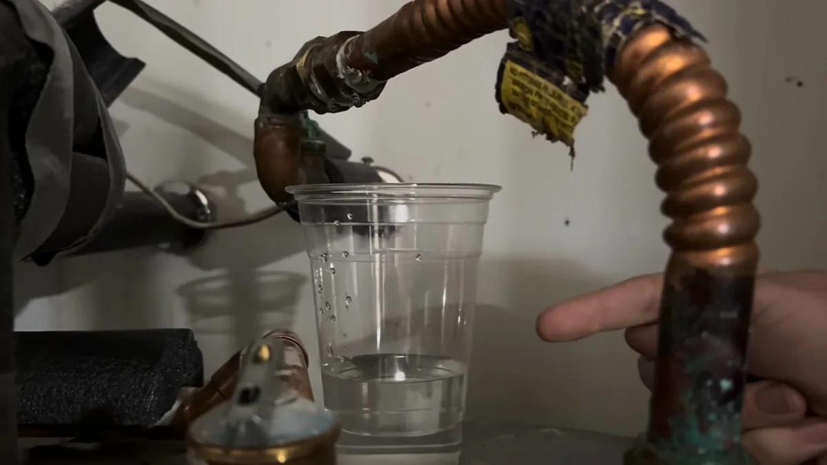 Green-stained copper pipe and a flexible connector at a water heater, with a clear plastic cup beneath and a finger pointing