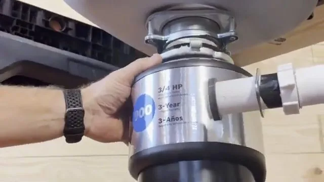 Hand gripping a stainless garbage disposal unit with the white discharge pipe attached beneath a sink