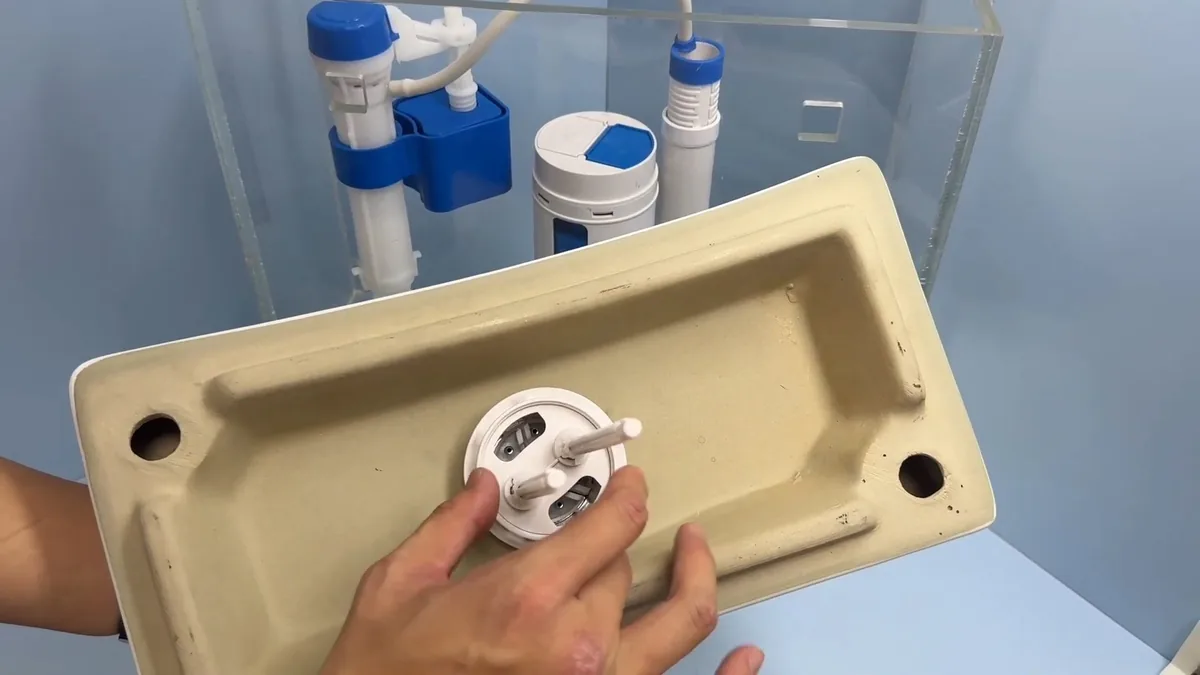 Hand holding a toilet tank lid upside down beside the open tank, showing the fill valve and flush valve inside