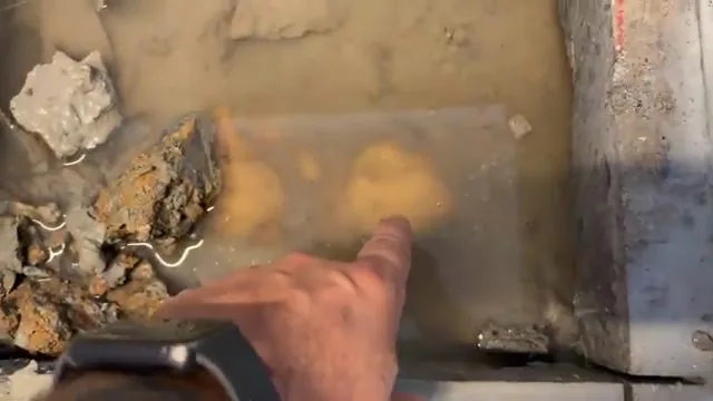 Hand pointing at brown water sitting in a broken concrete slab opening beside chunks of rubble