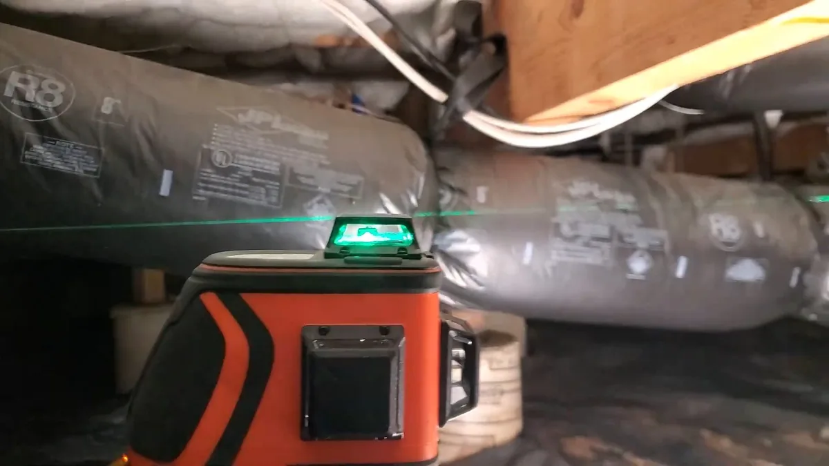 Orange laser level throwing a green line across wrapped ductwork and pipe in a crawlspace
