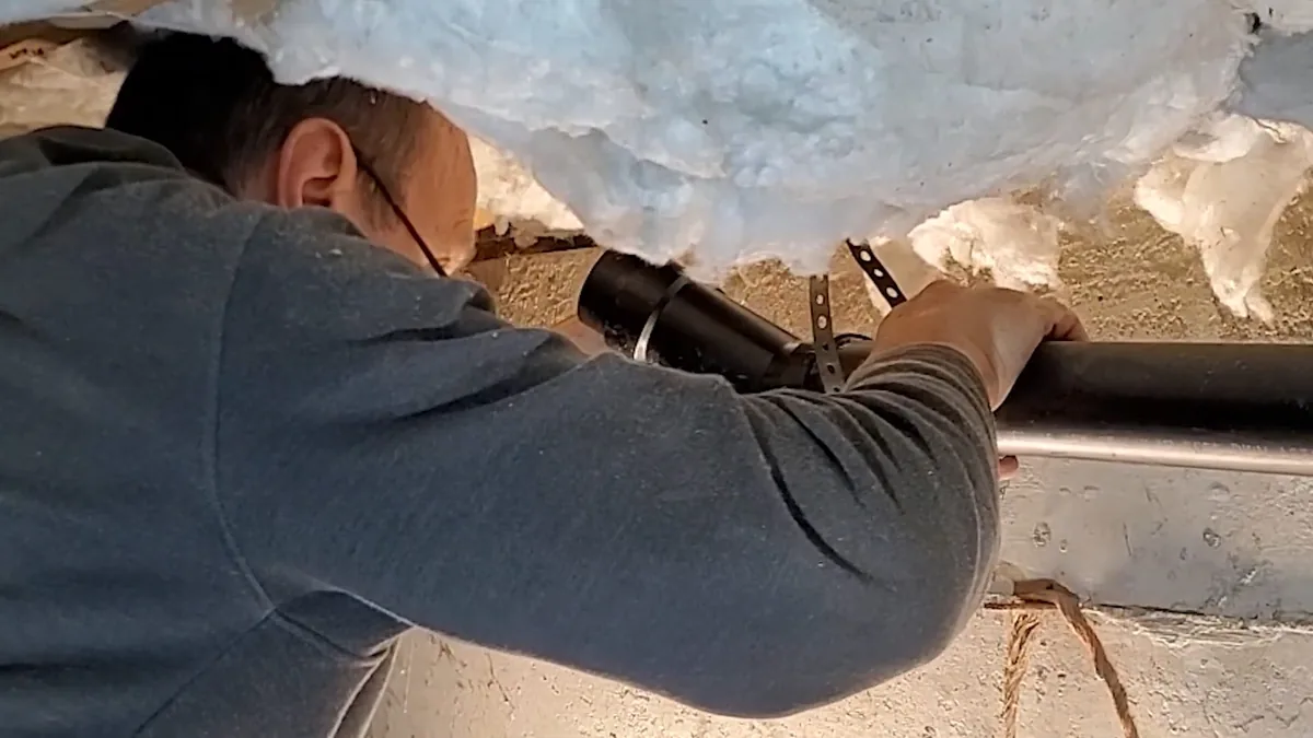 Man with glasses reaching up through blue insulation to check a black drain pipe and strap in a crawlspace