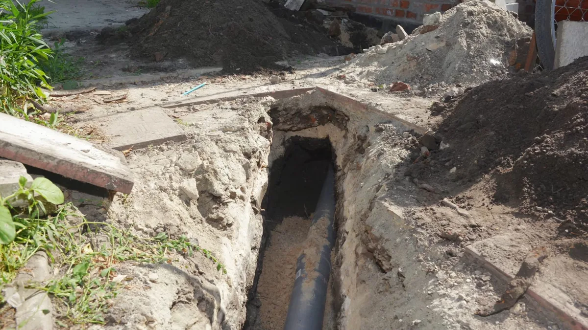 Narrow open trench beside a house wall, with a black pipe lying in the bottom and dirt piled alongside