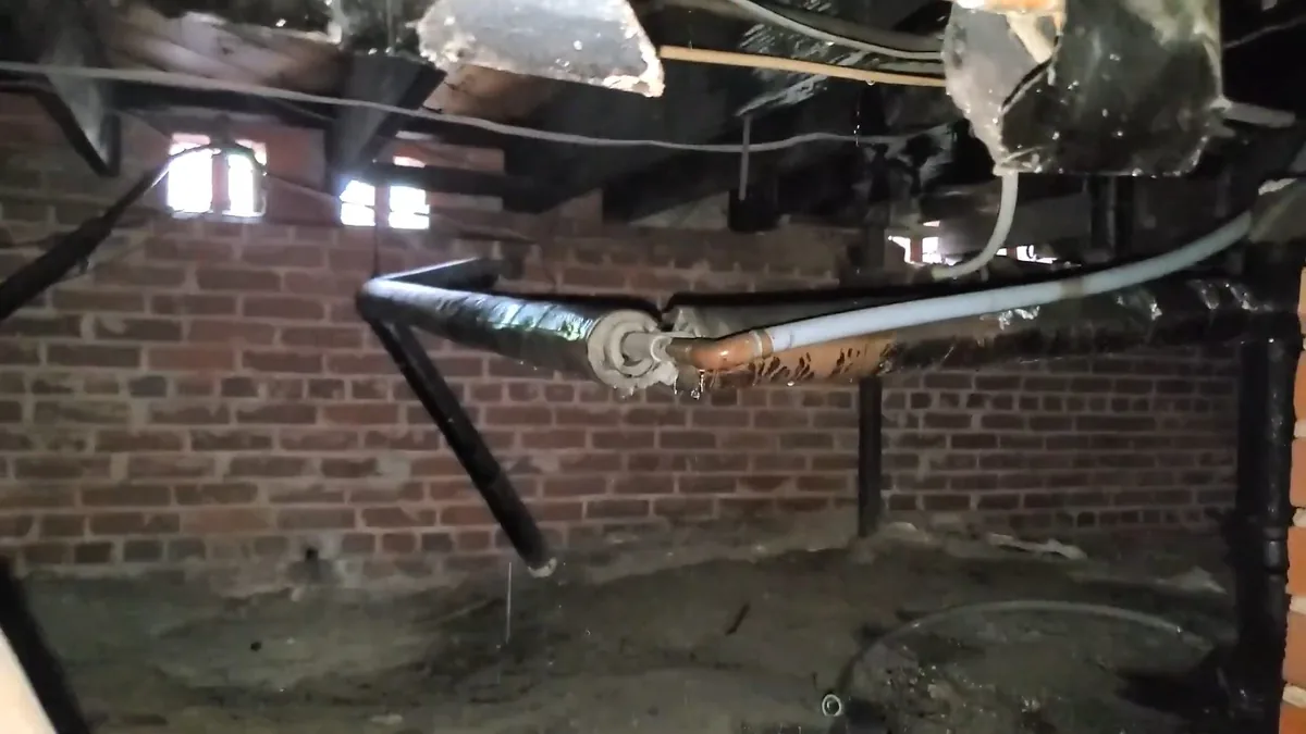 Black pipe and a copper line running along a red brick crawlspace wall beneath wood joists with loose insulation