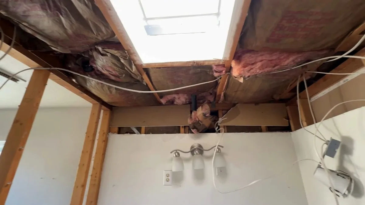 Opened ceiling above a white wall with a three-lamp vanity light, exposed pink insulation and a skylight well