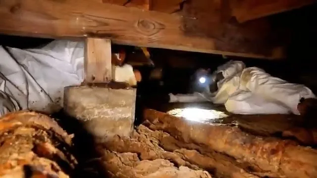 Person in a white protective suit with a headlamp crawling beneath floor joists beside a heavily rusted pipe