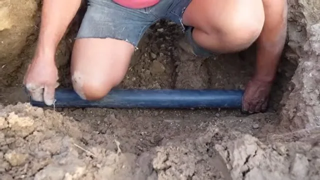 Person kneeling in a dirt trench, pressing a section of blue pipe into place