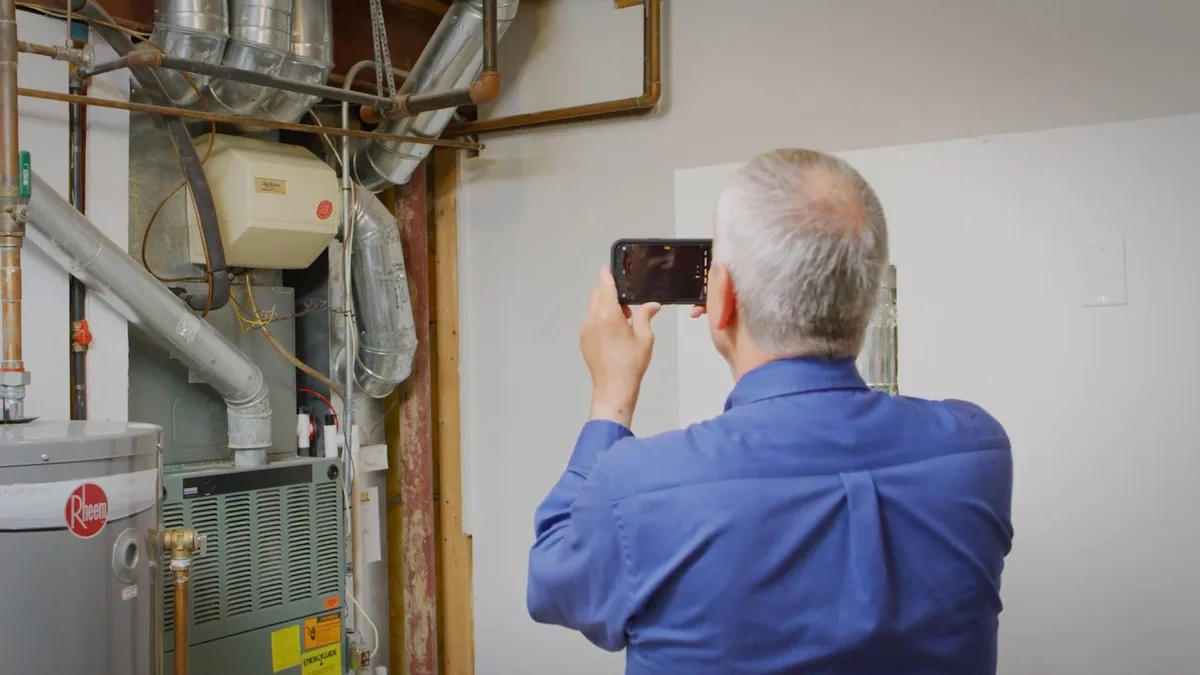 Person holding up a phone to photograph a water heater and the pipework around it