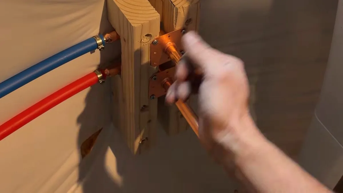 Hand reaching toward red and blue PEX lines connected to copper stub-outs on a wood block inside a bathroom wall