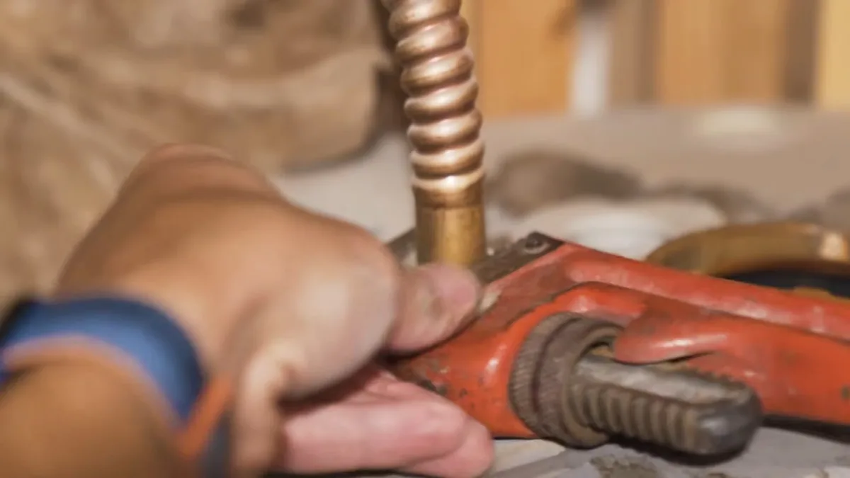 Hand gripping a red pipe wrench on the brass fitting of a corrugated flexible water connector at the top of a heater