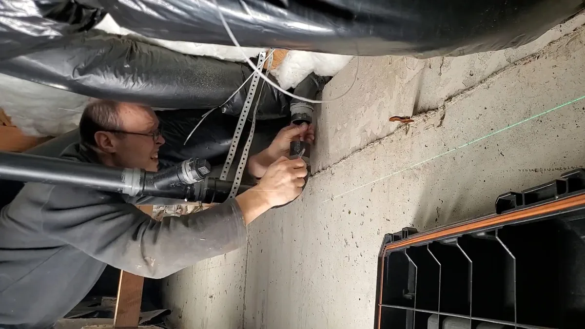Man leaning into a low space to fasten a drain pipe to a concrete wall beside wrapped ductwork, with a green laser line across the wall