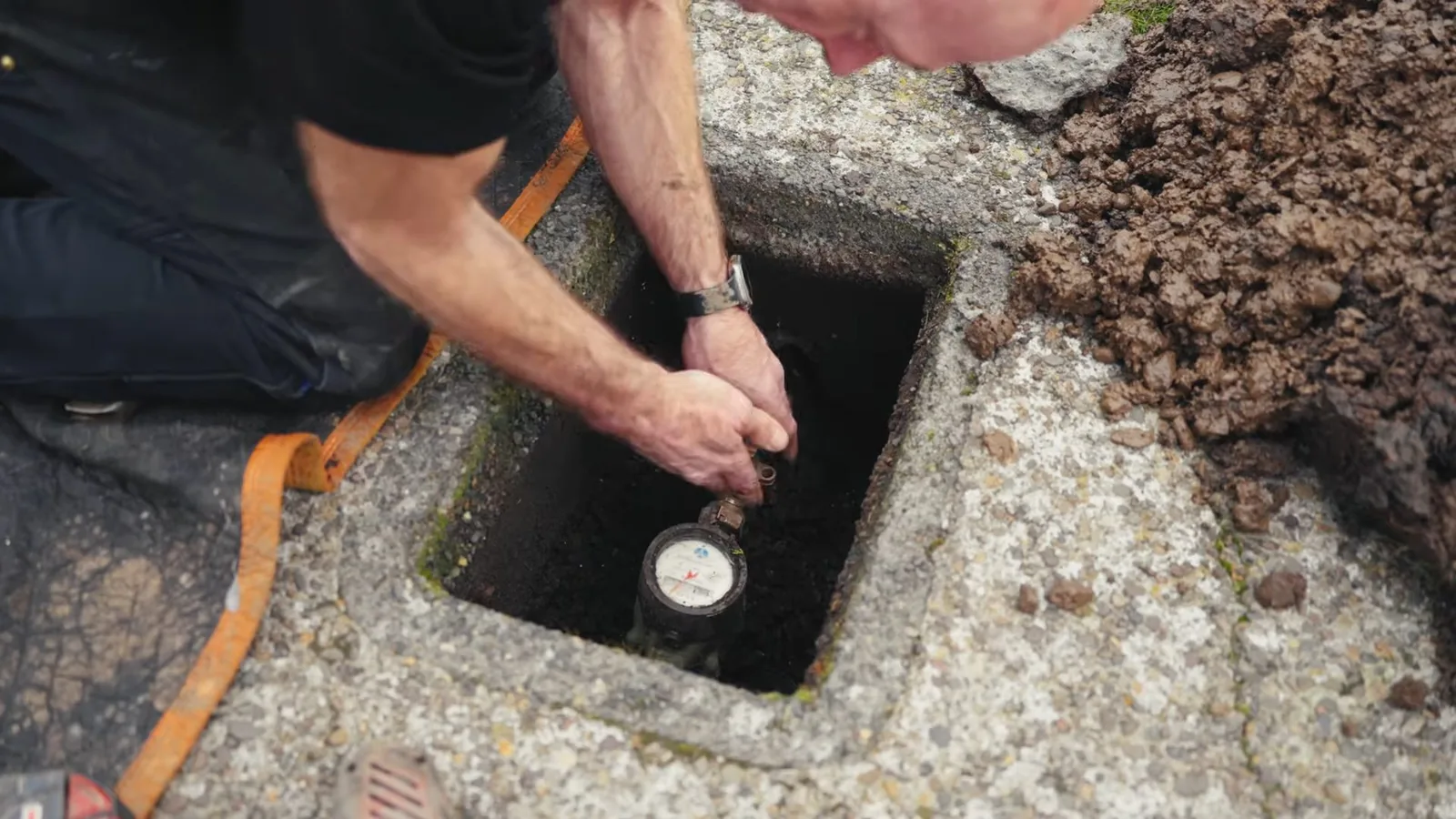 Plumber kneeling beside an open ground box to read the water meter inside it