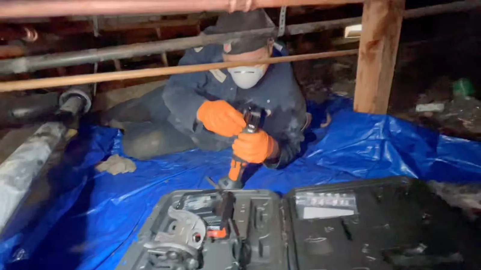Plumber in a dust mask kneeling on a blue tarp in a crawlspace, drilling near copper pipes