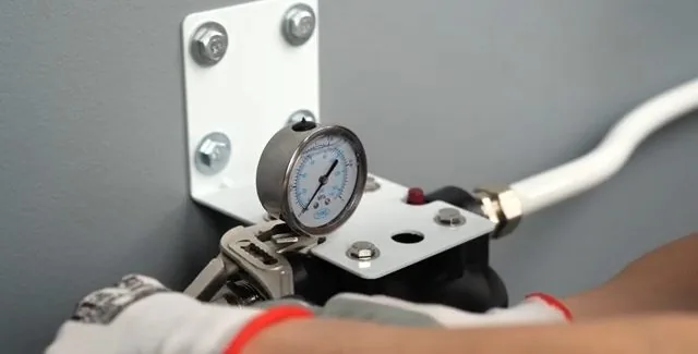 Gloved hand steadying a pressure gauge mounted on a white metal bracket beside a white supply line