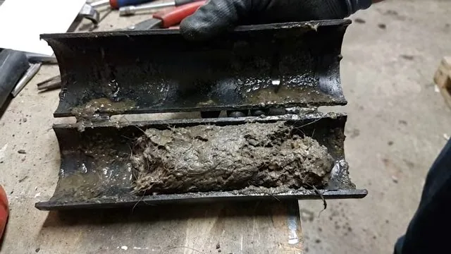 Metal drain pipe cut in half on a workbench, packed with grey sludge along its length