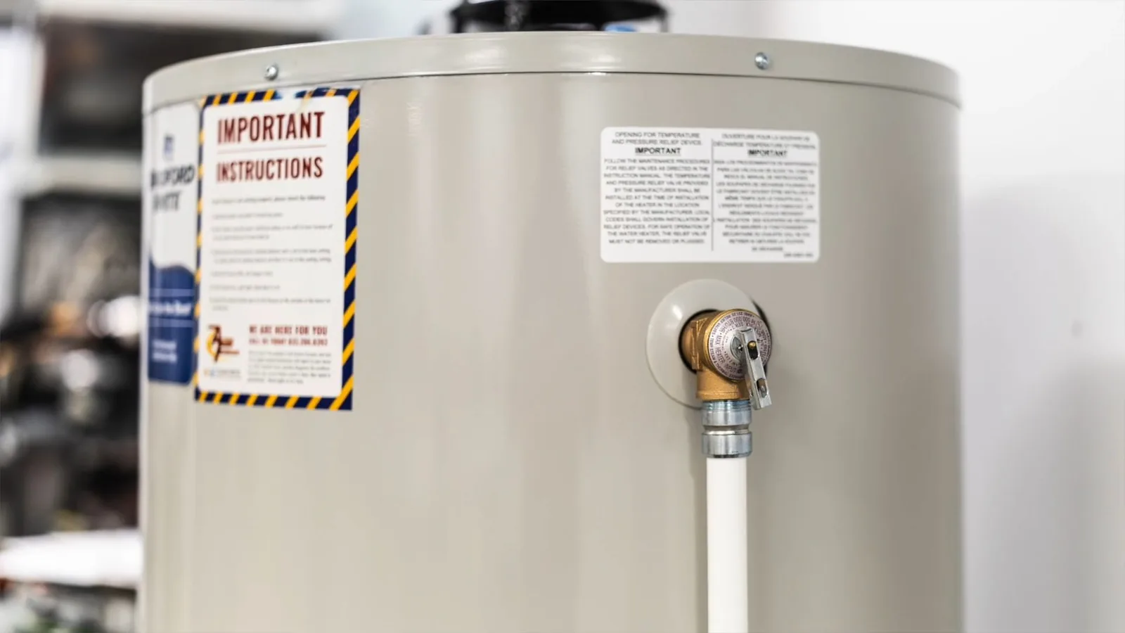Side of a new white water heater tank with a warning label and a white supply line on its connection