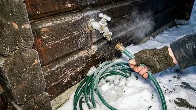 Gloved hand holding a green garden hose beside a steaming brass faucet on a log wall in the snow