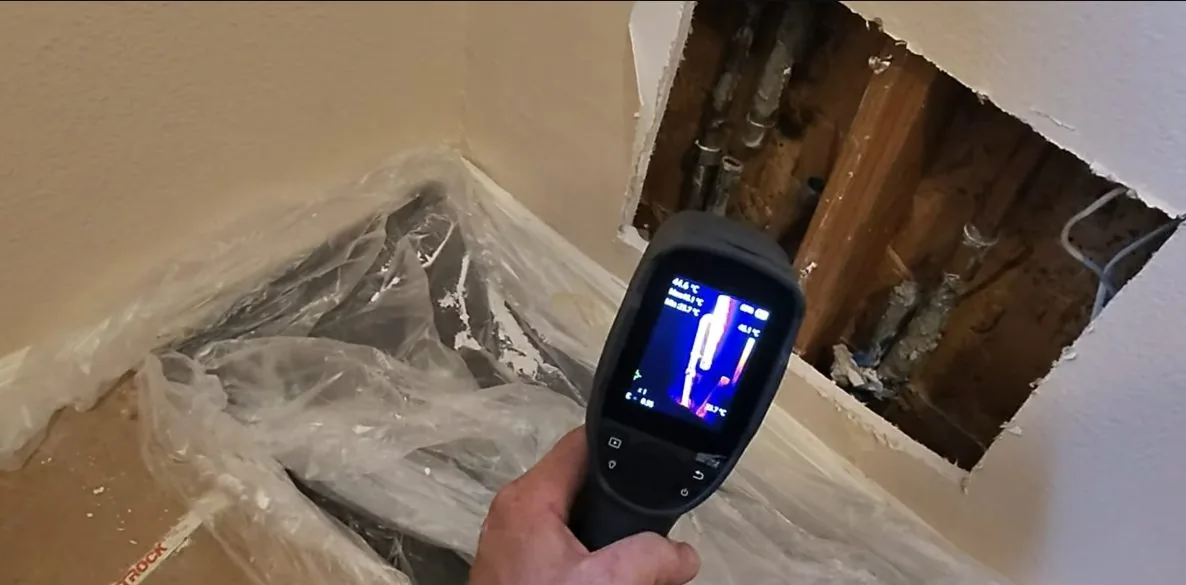 Hand holding a thermal imaging camera pointed at an opened wall and a plastic-covered floor corner