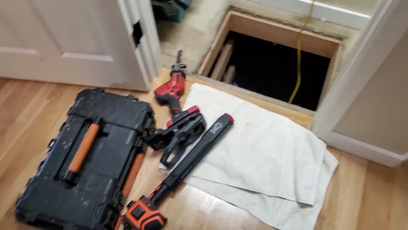 Black tool case, drill and pipe wrench laid on a wood floor beside an open crawlspace hatch
