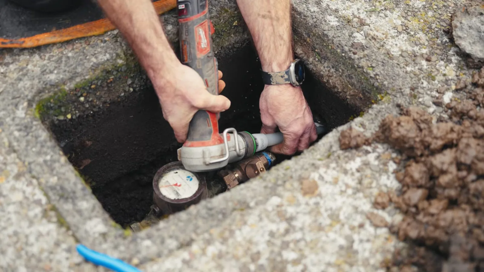 Hands holding a power tool over a water meter and shutoff inside a stone-lined meter box