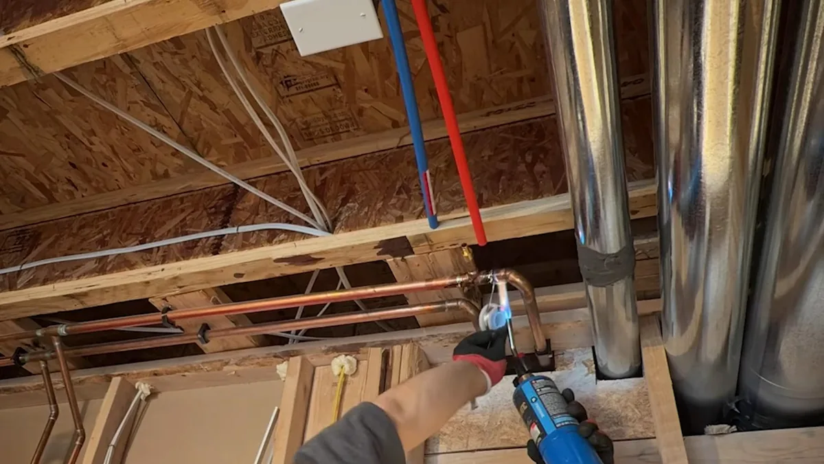Hand in a red glove holding a torch to a copper joint among red and blue PEX lines and metal ducts under a basement ceiling