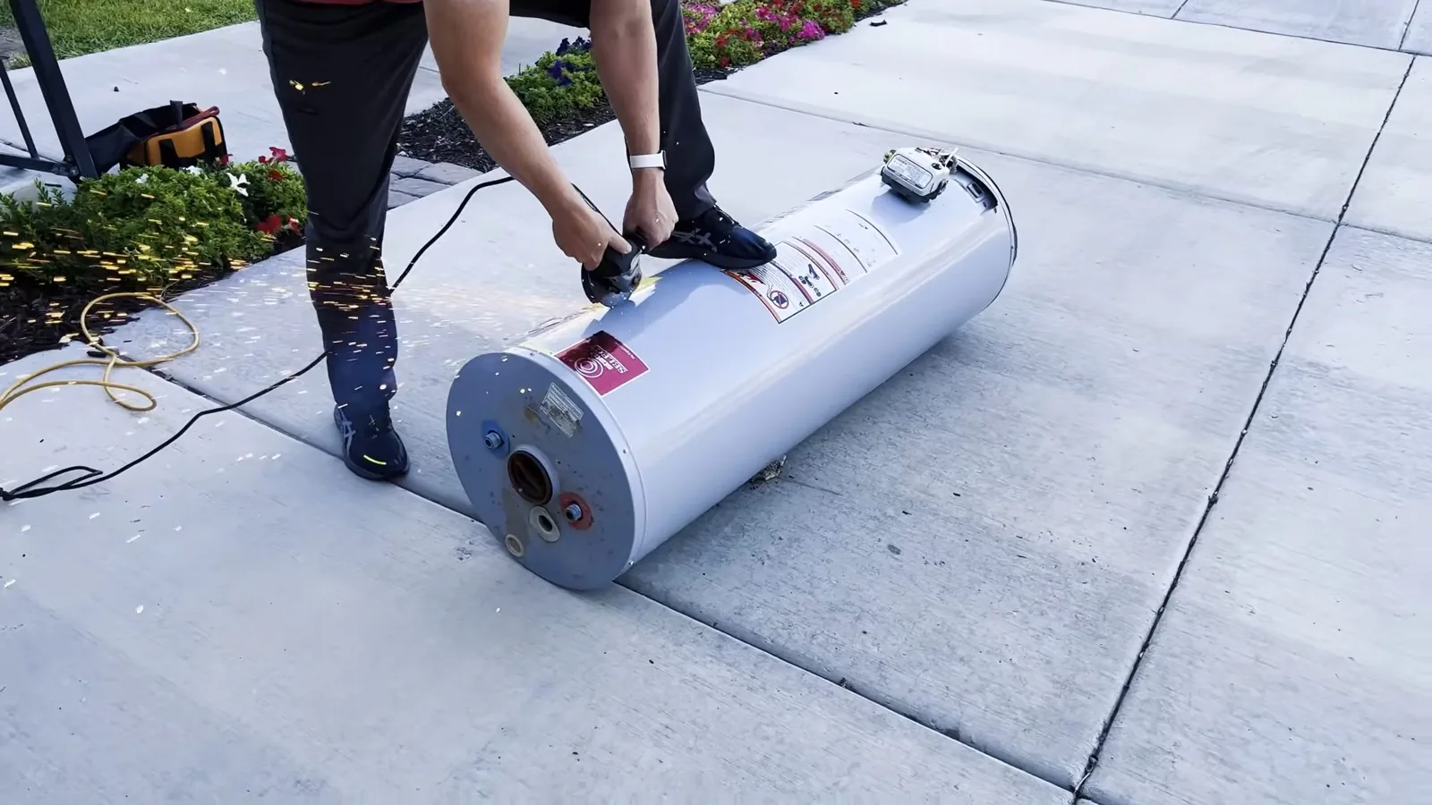 Two people carrying a new water heater tank lying on its side across a paved driveway