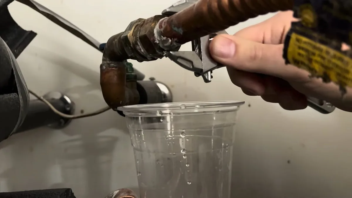 Hand loosening a heater fitting with water running into a clear plastic cup below it