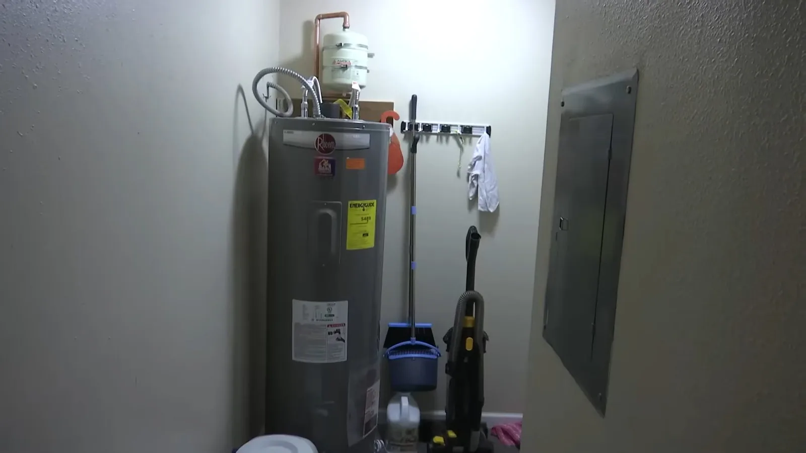 Electric water heater standing in a bare utility room with a panel on the wall beside it