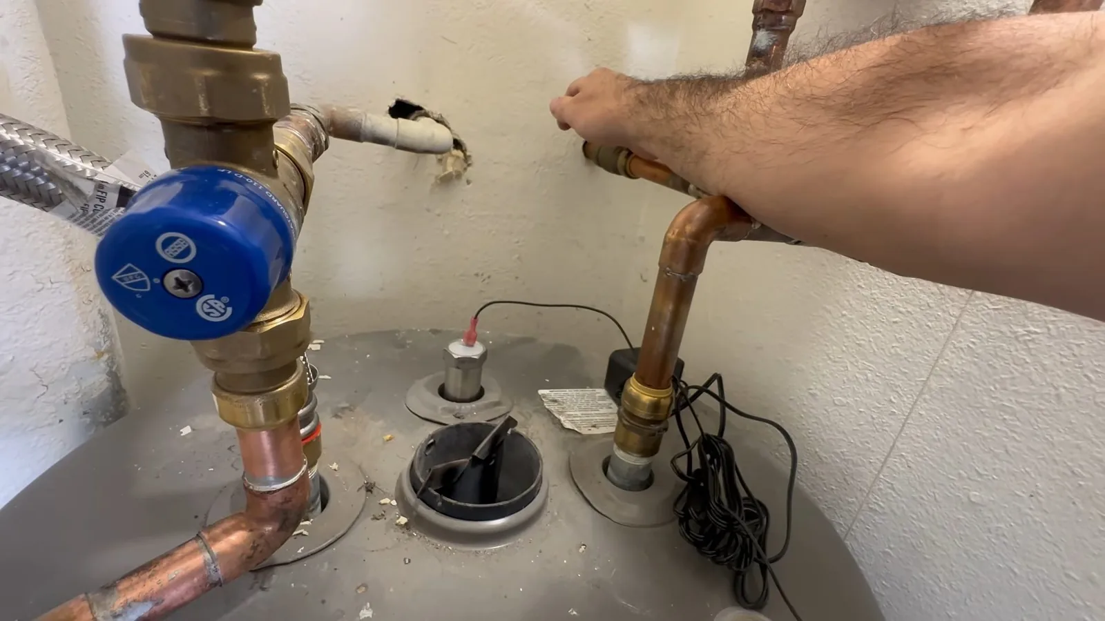 Top of a water heater with a blue valve, a flexible hose and copper lines, and a hand adjusting a fitting