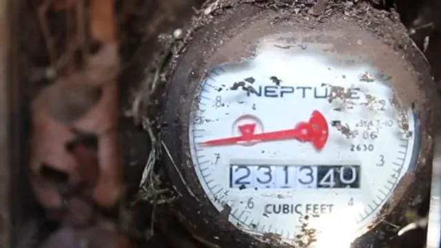 Close view of a water meter dial with a red sweep hand and a cubic feet readout, dusty from its box