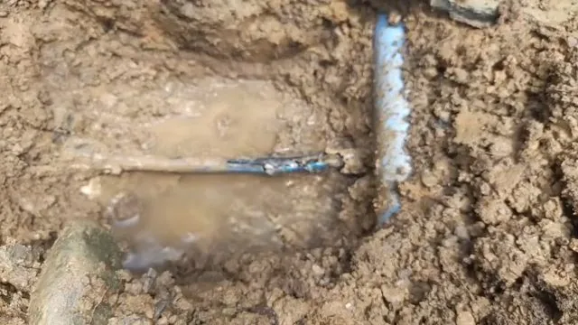 Muddy excavation with water pooling around a blue plastic pipe where it has been cut