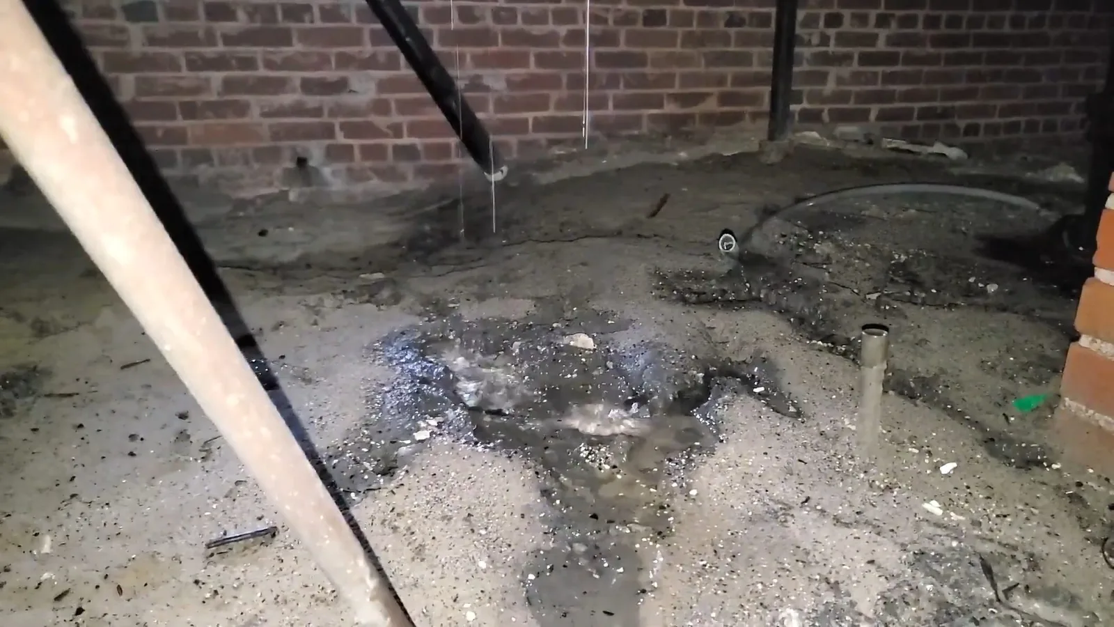 Wet, dark basement floor against a brick wall with water seeping near a pipe stub