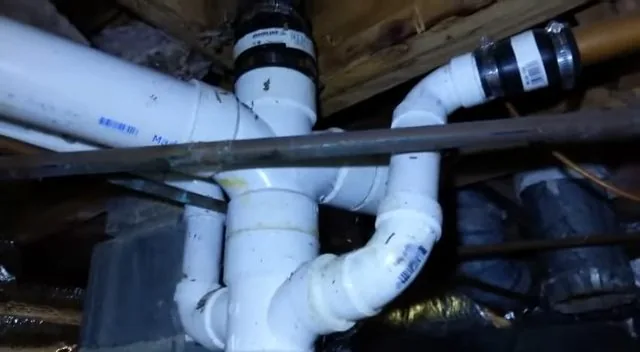 White PVC drain junction with rubber couplings in a tight crawlspace under wood joists